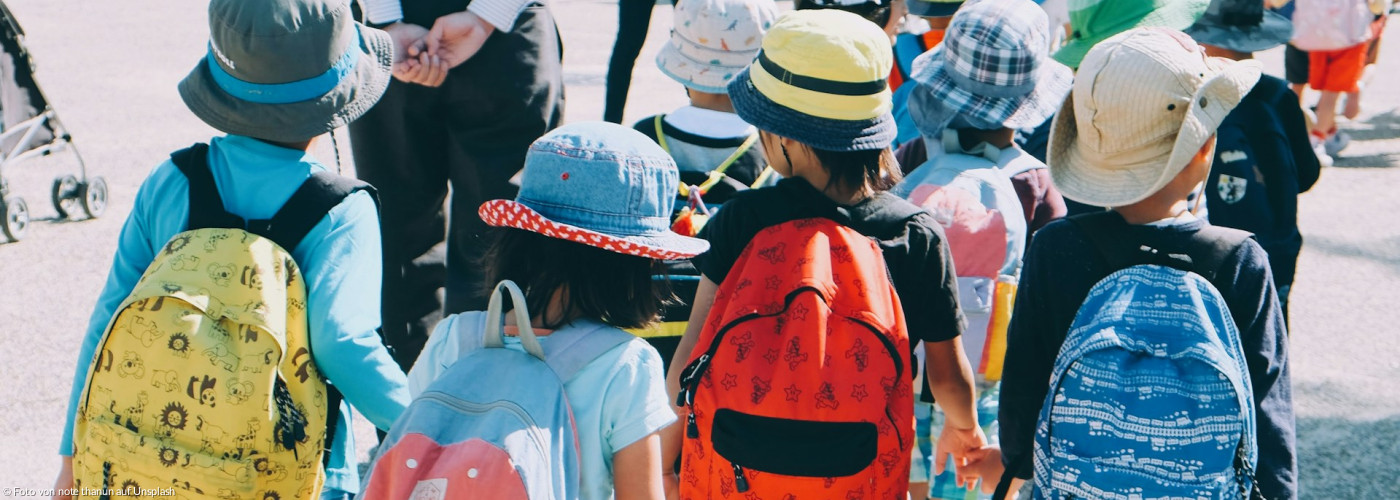 Gruppe von Kindern, die weiße und orangefarbene Rucksäcke tragen und tagsüber auf grauem Betonpflaster spazieren gehen"
