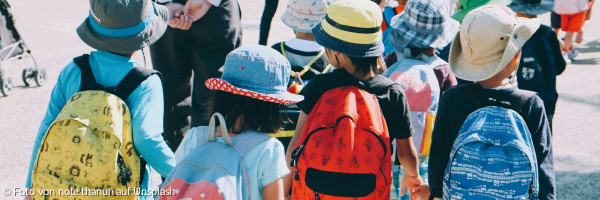 Gruppe von Kindern, die weiße und orangefarbene Rucksäcke tragen und tagsüber auf grauem Betonpflaster spazieren gehen"